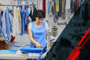 Woman in a blue apron ironing clothes at a garment ironing station amid hanging garments in a laundry setting.