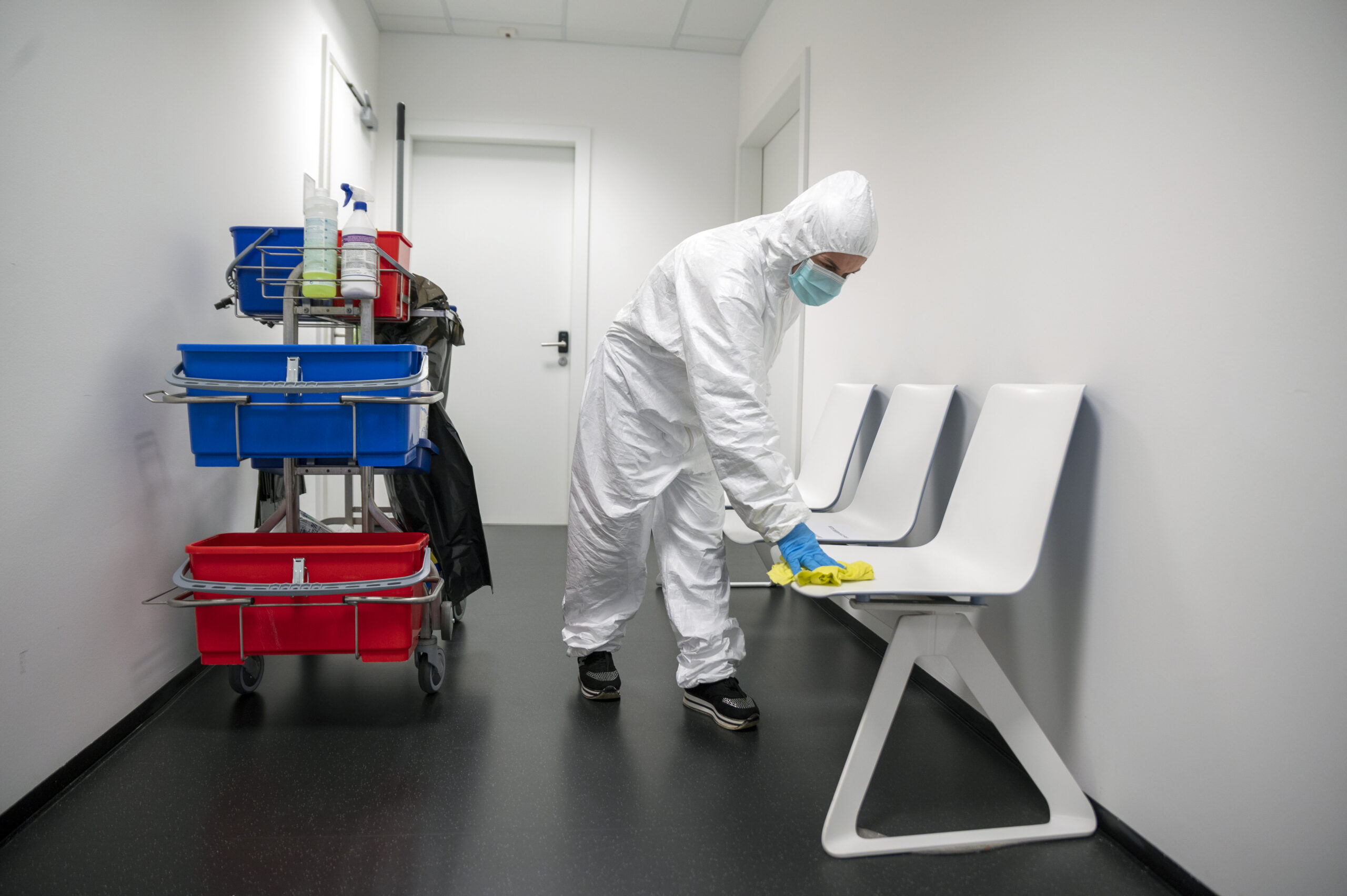 Person in a white hazmat suit and mask disinfecting a hospital hallway with a cleaning cart nearby.