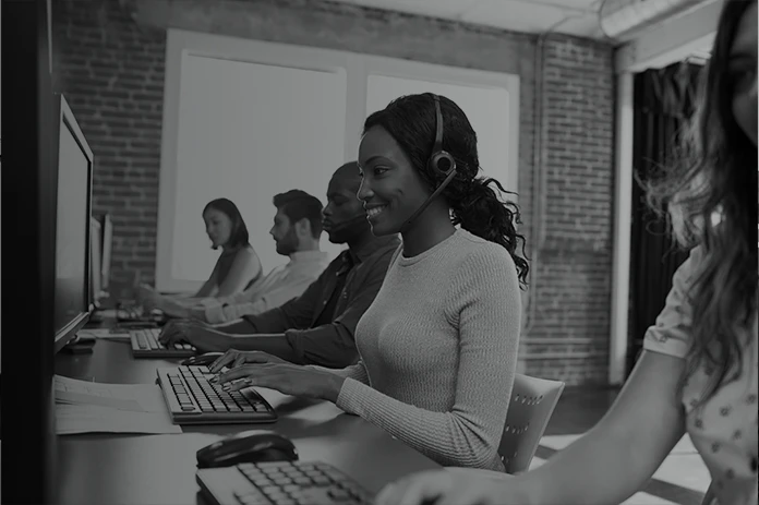 Woman wearing a headset sits at a computer in a row of call center agents, smiling as she types.